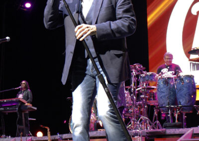 A man in sunglasses, a dark blazer, and jeans sings into a microphone on stage, with band members playing instruments in the background and colorful lights and a large graphic on the screen behind them.