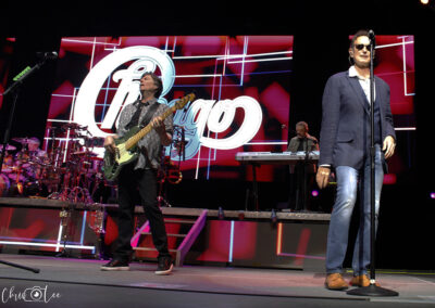A band performs on stage with a large Chicago logo displayed on a screen behind them. One musician plays bass guitar while another, wearing sunglasses and a suit jacket, sings into a microphone. Instruments and lights are visible.