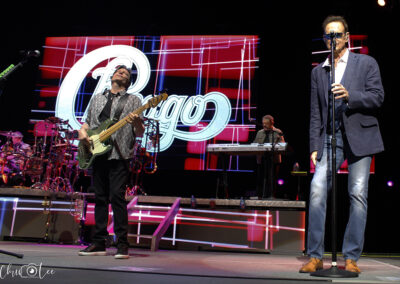 A rock band performs on stage with a large Chicago logo displayed on a screen in the background. One musician plays guitar while another sings into a microphone. Bright stage lights illuminate the scene.