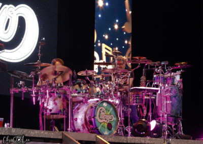 Two drummers perform on a brightly lit stage surrounded by multiple drums and cymbals, with a large illuminated Chicago band logo displayed in the background. The scene is vibrant with pink and purple lighting.