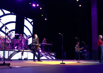 A band performs on a brightly lit stage with musicians playing guitar, saxophone, keyboard, and trumpet. Colorful lights and abstract designs illuminate large screens behind them.