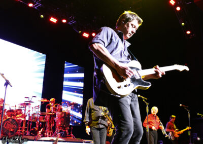 A band performs on stage under colorful lights. The guitarist in the foreground plays energetically, while other band members play instruments, including drums and keyboards, with a bright digital backdrop behind them.