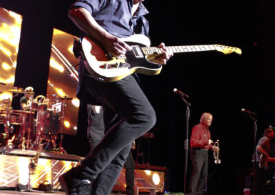 A guitarist plays energetically on stage, mid-jump, with a band performing in the background under bright stage lights. Other musicians, including a brass player, stand nearby, with vibrant screens behind them.
