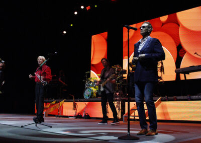 A band performs on stage with colorful orange and yellow lights in the background. Several musicians stand with instruments, a singer is at the front, and a keyboardist is silhouetted at the back.