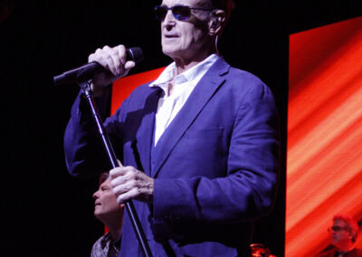 A man in a blue suit and sunglasses sings into a microphone on stage, with musicians and red-orange light panels in the background.