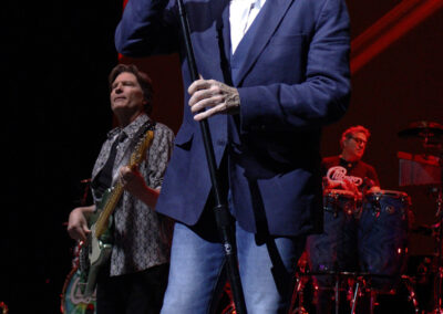 A man in a blue blazer, white shirt, and sunglasses sings into a microphone on stage; a guitarist and a drummer perform behind him under stage lights.