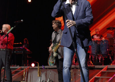 A male singer in sunglasses and a blazer holds a microphone on stage, with band members playing instruments in the background under colorful stage lights.