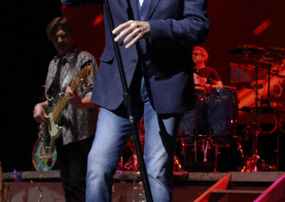 A man in a blue blazer, white shirt, and jeans sings into a microphone on stage. Musicians play instruments behind him, and stage lights create a dramatic atmosphere.