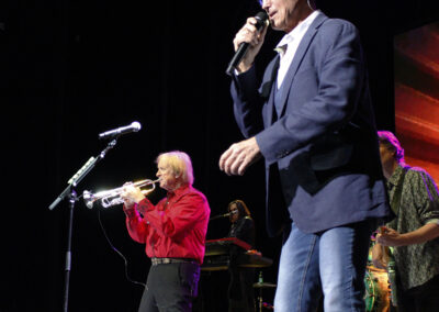 A singer in sunglasses and a blue blazer performs with a microphone while a trumpet player in a red shirt plays beside him on stage. Musicians and instruments are visible in the background under colorful stage lighting.