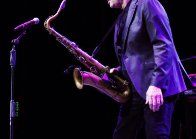 A man in a black suit plays a saxophone on stage under bright lights, standing near a microphone with a dark background.