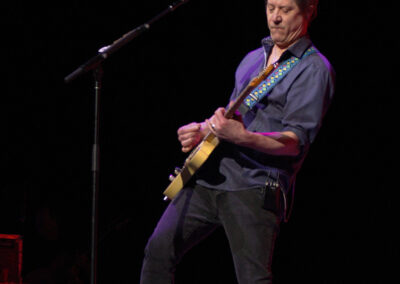 A musician in a blue shirt and black jeans plays an electric guitar on stage, leaning into the music. The background is dark and the stage is lit, with a microphone stand in front of the performer.
