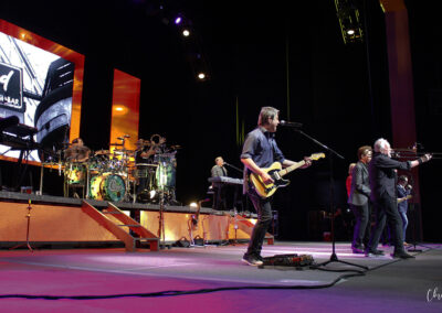 A band performs on stage with various instruments including guitar, drums, keyboard, and trombone. Bright lights and large screens with graphics illuminate the background. The musicians are actively engaged in their performance.