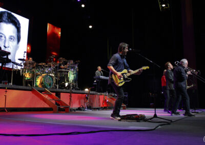 A band performs on a brightly lit stage with multiple musicians, including a guitarist, trombone player, and drummer. Large screens behind them display black-and-white faces. Stage lights and instruments are visible.