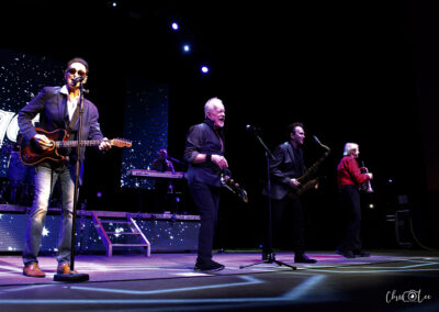Four musicians perform on stage under colorful lights; one plays guitar and sings, others play brass instruments and keyboard, with a backdrop of stars and a sparkling band logo.