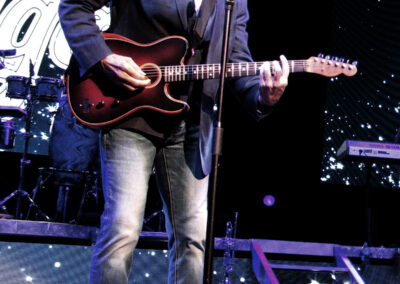 A man in sunglasses, a blazer, jeans, and brown shoes plays an electric guitar and sings into a microphone on stage, with a starry backdrop and bright stage lights behind him.