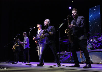 A band performs live on stage with guitars, a saxophone, microphones, and drums, backed by a starry, illuminated screen. The musicians are mid-song, engaging with the audience under stage lighting.