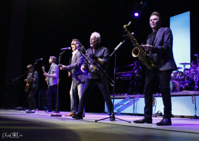 A band performs energetically on stage, with six musicians playing guitars, saxophones, and singing into microphones under bright stage lights. The background includes a drum set and illuminated blue screens.
