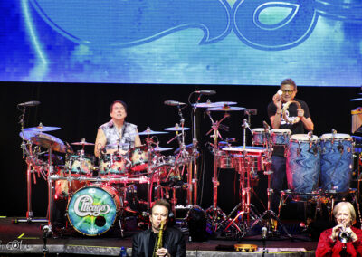 A band performs on stage with two drummers surrounded by drums and percussion, one playing a drum set with a Chicago logo. Other band members play trumpet and saxophone, with a blue-lit background behind them.