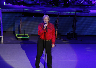An older man wearing a red shirt and black pants sings into a microphone on stage, with musical instruments and drum set labeled “Chicago” visible in the background under concert lighting.