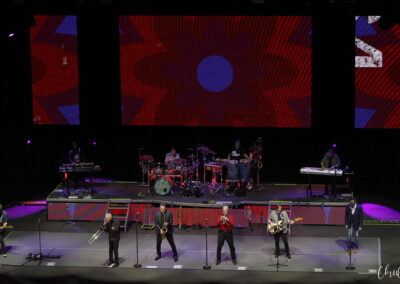 A band of nine musicians performs on stage with various instruments, including brass, guitars, keyboards, and drums, in front of three large screens displaying colorful abstract flower patterns.