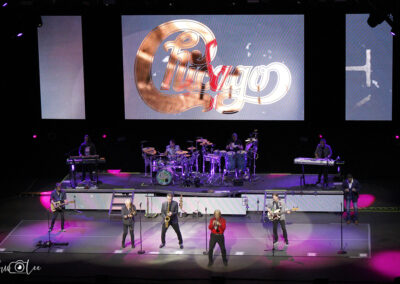 A band performs on stage with a large screen behind them displaying the Chicago logo. The stage is lit with purple and pink lights, and multiple musicians play instruments including guitars, drums, and horns.