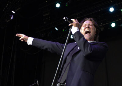 A man in a dark suit passionately sings into a microphone on stage, eyes closed and mouth open wide, with one arm extended and pointing, against a dark background with stage lights above.