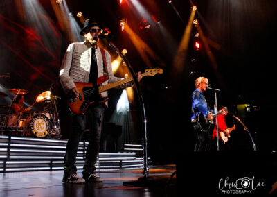 A rock band performs onstage under colorful lights; the bassist sings into a microphone, the drummer plays in the background, and two guitarists perform in front of a cheering audience. Chris Lee Photography watermark is visible.