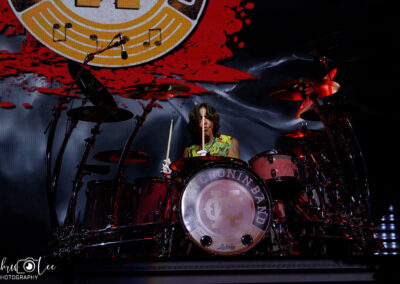 A drummer plays a red drum set on stage under dramatic lighting, with a large “REO Speedwagon” logo displayed in the background. The drum kit features prominent cymbals and the band’s name on the bass drum.