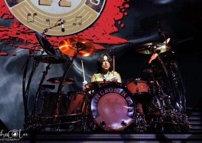 A drummer performs energetically on stage, surrounded by a large drum kit. Colorful lights illuminate the scene, and a circular logo with a red background appears behind the drummer. The drum reads LATIN CROWN BAND.