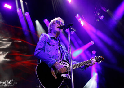 A musician wearing glasses and a denim jacket plays an acoustic guitar and sings into a microphone on stage, surrounded by dramatic purple and white stage lights.