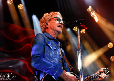 A musician with curly blond hair and glasses plays guitar and sings into a microphone on stage, surrounded by colorful lights and dramatic lighting effects.