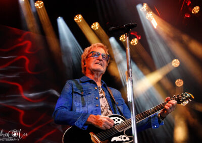 A man wearing sunglasses and a denim jacket plays an acoustic guitar and sings into a microphone on stage, with colorful stage lights and a red patterned background behind him.