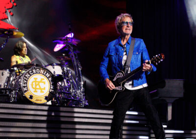 A guitarist in a blue jacket and sunglasses performs on stage, smiling, while a drummer plays a white drum kit in the background. Bright stage lights and musical equipment are visible.