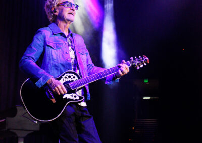 A musician wearing glasses and a denim jacket plays an acoustic guitar on stage under purple and white spotlights, with a piano and steps in the background.