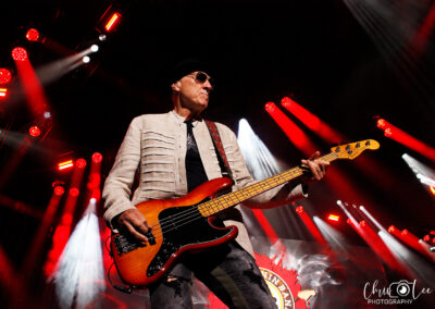 A musician wearing sunglasses and a light-colored jacket plays an electric bass guitar on stage, surrounded by dramatic red and white stage lights. A band logo is visible in the background.