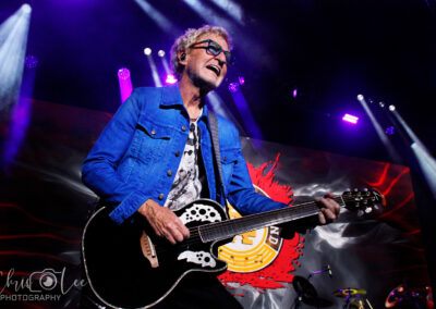 A musician in a blue jacket and glasses plays a black acoustic guitar on stage, with colorful lights and a vibrant background behind him. The image is credited to Chris Lee Photography.
