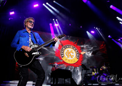A guitarist in a blue jacket performs energetically on stage with a band. Bright purple stage lights shine above them, and a large screen in the background displays the Kevin Cronin Band logo.