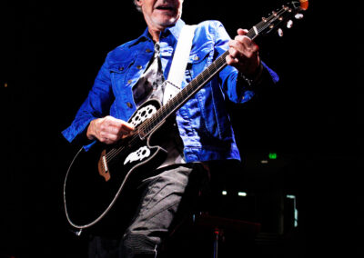 A musician with blond hair wearing sunglasses and a blue jacket plays a black acoustic guitar on stage under bright lights, with a dark background.