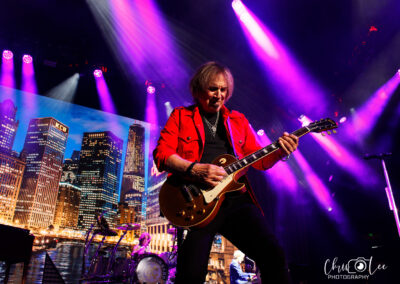 A guitarist in a red jacket plays on stage under vibrant purple lights, with a city skyline backdrop and a drummer performing in the background. The stage has vivid colors and dynamic lighting.