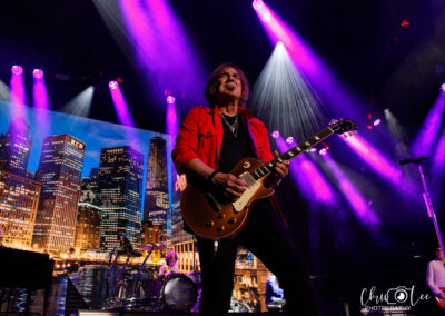 A guitarist in a red shirt plays an electric guitar onstage under purple lights, with a drummer in the background and a city skyline image displayed behind them.