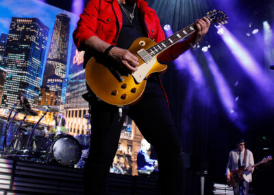 A guitarist in a red jacket plays an electric guitar on stage under dramatic purple and white lights, with a cityscape displayed on a screen in the background and other band members performing nearby.
