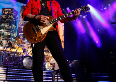 A guitarist in a red jacket performs energetically on stage under purple lights, with a cityscape backdrop and a drummer visible in the background. The atmosphere is vibrant and lively.