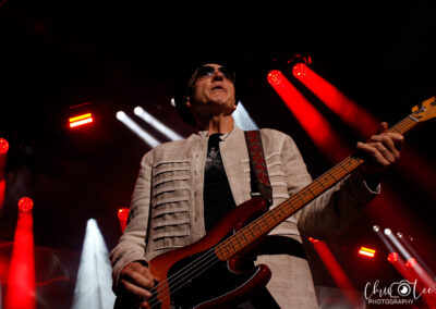 A musician wearing sunglasses, a hat, and a light-colored jacket plays an electric bass guitar on stage, surrounded by dramatic red and white stage lights. The photo has a watermark that reads Chris Lee Photography.