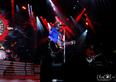 A rock band performs on stage under red and blue lights; the lead singer plays guitar and sings at the microphone, with a drummer and guitarist in the background, and a large band logo displayed on a screen.