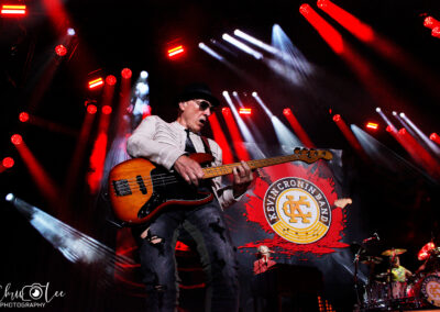A musician wearing sunglasses and a hat plays an electric bass guitar on stage, with bright red and white lights shining. The backdrop displays the Kevin Cronin Band logo. Drums and another musician are partially visible.