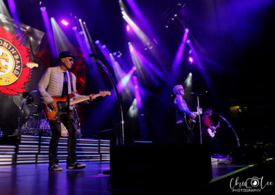 A rock band performs on a brightly lit stage with purple lights. Three musicians play guitar and sing, while a drummer is visible in the back. The bands logo is displayed on a large screen behind them.