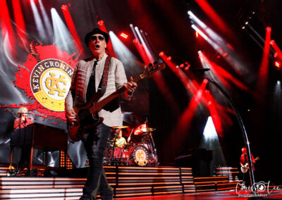A rock band performs on a brightly lit stage with red and white lights. The bassist stands in front, singing into a microphone. Behind him, a drummer and keyboardist play, with the bands logo Kevin Cronin displayed on a large screen.