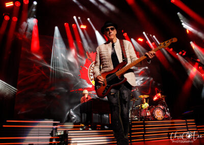 A musician in a white jacket and hat plays bass guitar on stage with dramatic red lighting. A drummer and keyboardist are visible in the background, and a large band logo is displayed behind them.