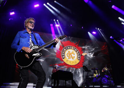 A musician in a blue jacket plays guitar on stage, with a large screen behind showing Kevin Cronin Band logo. A keyboardist and drummer perform under purple stage lights.