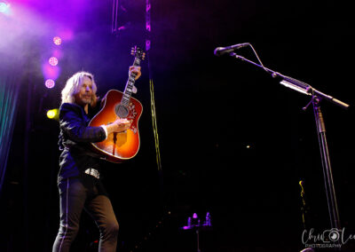A musician with long hair holds up an acoustic guitar on stage under colorful purple and yellow lights, with a microphone stand and water bottles visible nearby.